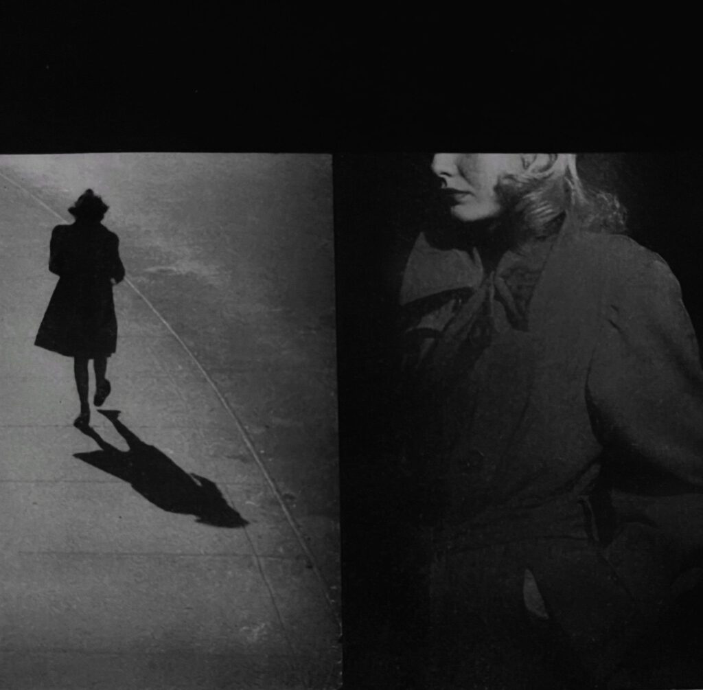 Black and white photo of a the back of a woman walking down the street while another woman watches